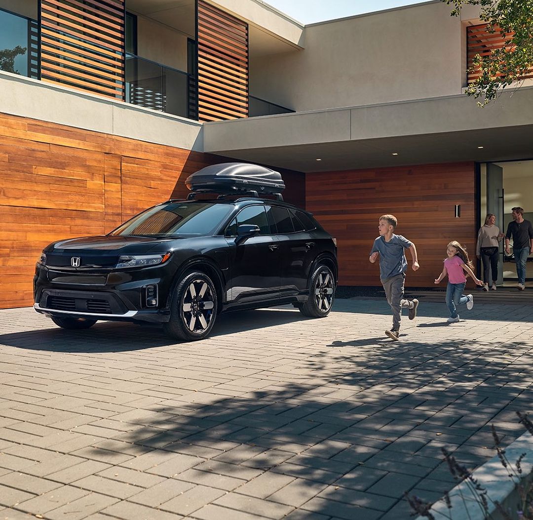 Black Honda Prologue SUV with roof cargo carrier parked outside a modern house as children run towards it, family watching in the background.