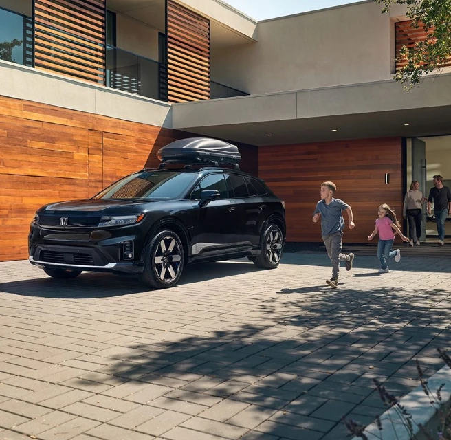 Black Honda Prologue SUV with roof cargo carrier parked outside a modern house as children run towards it, family watching in the background.