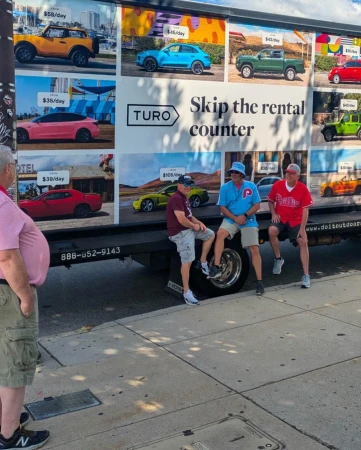 Three men sit on a truck advertising Turo car rentals, showcasing a variety of cars with pricing. One man stands on the sidewalk nearby.
