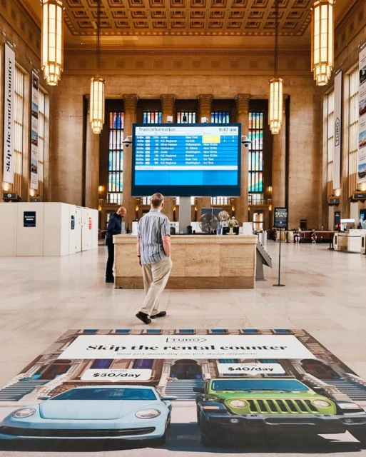 An adult male stands in a train station, looking at the train schedule. An advertisement for a car rental company is on the floor.