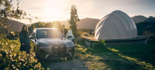 Luxury silver BMW iX electric SUV parked outdoors with a man and woman standing nearby, set against a natural backdrop with dome structure.