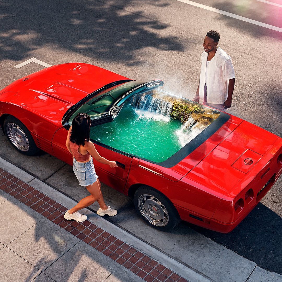 Overhead shot of a red convertible with two people, and an interior waterfall scene, parked on a city street with dappled light.