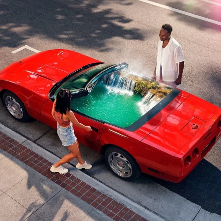 Overhead shot of a red convertible with two people, and an interior waterfall scene, parked on a city street with dappled light.