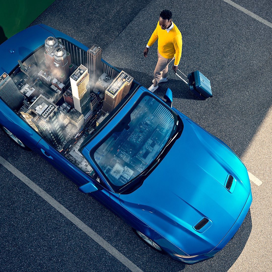 Aerial view: a blue convertible car filled with city buildings and a Black man in yellow sweater pulling a suitcase.