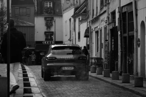 Monochrome street view of a luxury crossover car driving away down a cobblestone European alleyway with a pedestrian walking past in the foreground.