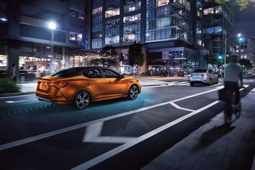 An orange Nissan Sentra sedan drives at night on a city street with digital safety assistance shown, other vehicles in background.