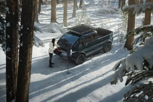 Woman accessing dark green electric truck in a snowy forest with ski poles, showcasing a blend of vehicle design and winter outdoor lifestyle.