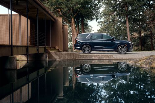 Dark blue SUV parks near modern home, reflected in still pond water, surrounded by trees under an overcast sky in an automotive shot.