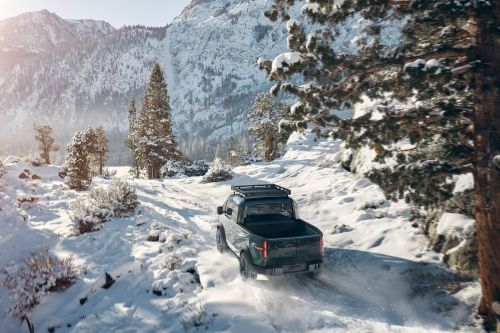 Gray pickup truck drives on snowy road in mountain landscape; evergreens and mountain peak under sunny skies in automotive shot.