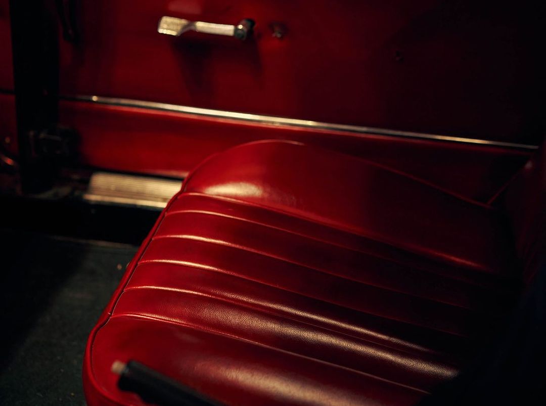 Close-up of a vintage car's interior featuring a glossy red leather seat with ribbed details and a chrome door handle.
