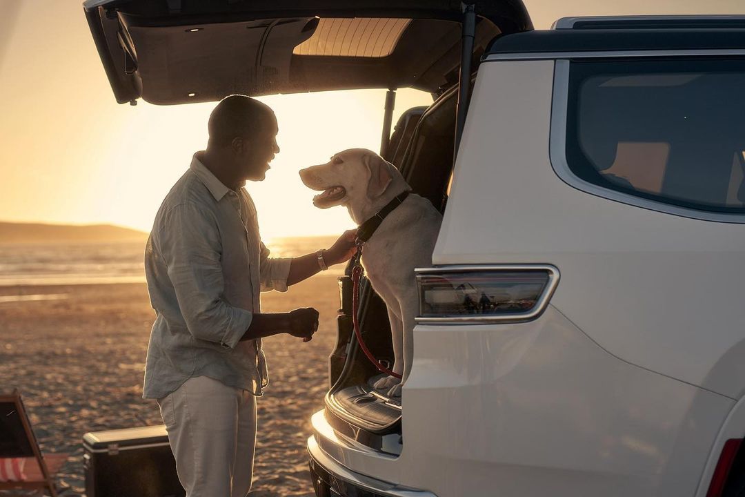 A man and his Labrador Retriever dog at the back of a car, enjoying the beach at sunset. A warm and inviting scene.