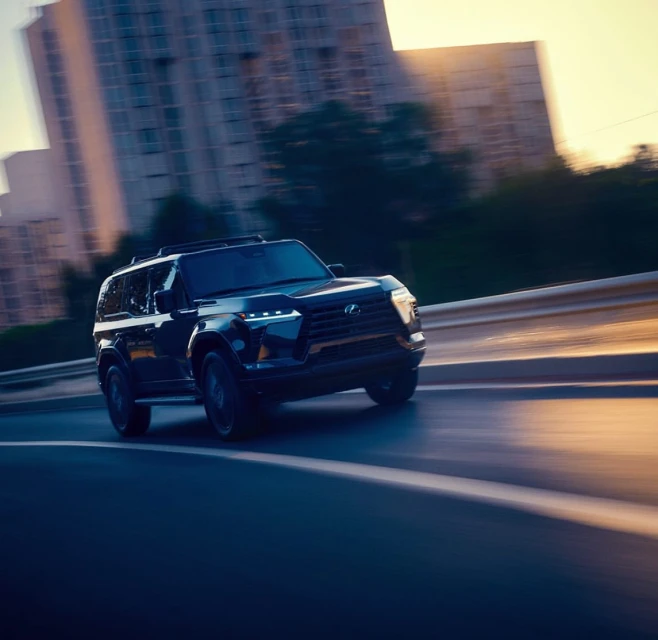 A dark SUV vehicle drives on an asphalt road in an urban setting with blurred motion suggesting speed.