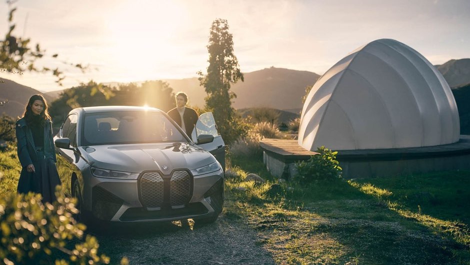 A gray BMW electric SUV is parked outdoors with a woman standing nearby and a man opening the driver's side door against a mountain backdrop.