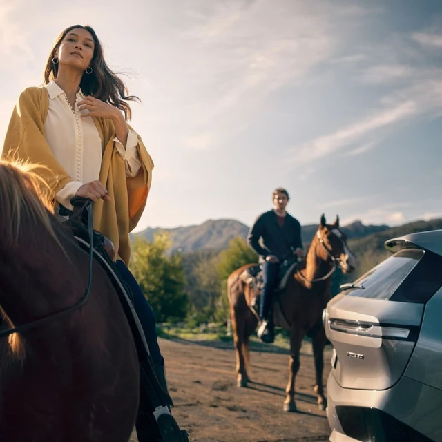 Woman in yellow shawl riding horse alongside man and modern SUV in a mountain landscape on a sunny day in an outdoor setting.