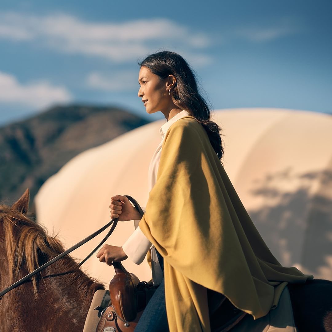 Woman riding a horse in a desert landscape with a yurt in the background on a sunny day, wearing jeans and a mustard-colored poncho.