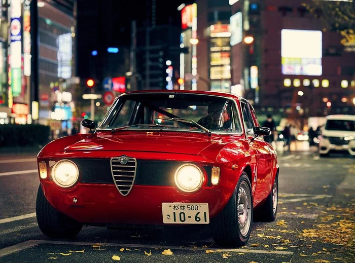 Vintage red Alfa Romeo classic car parked on a lit city street at night with glowing headlights and illuminated urban background