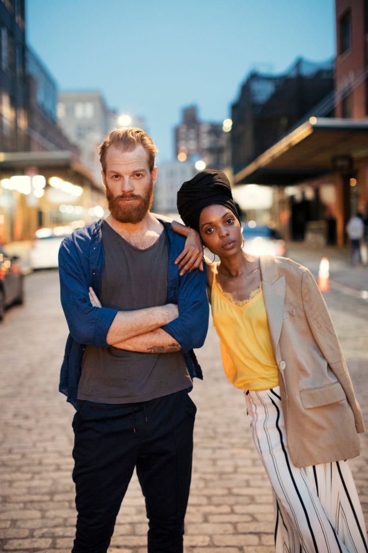 A confident couple poses outdoors against an urban background, with the red-haired man in a blue shirt and the woman in a yellow top and turban.