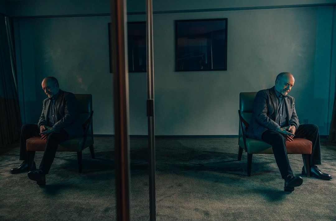 Man seated in an armchair reflected in a mirror inside a dimly lit room shot by Victoria Will