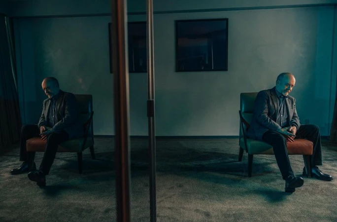 Man seated in an armchair reflected in a mirror inside a dimly lit room shot by Victoria Will