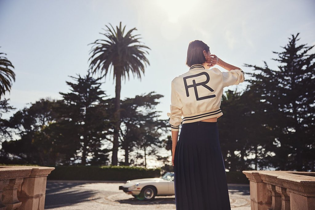 Woman in bomber jacket and long skirt stands in front of lush greenery and a vintage car on a sunny day in an outdoor setting.