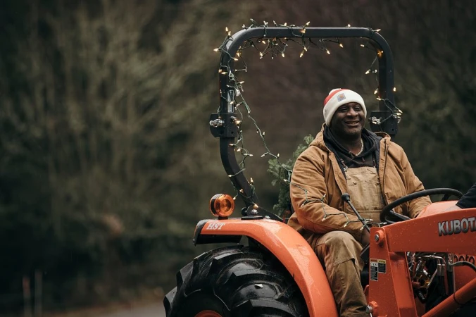Adult man wearing a Santa hat and overalls drives an orange Kubota tractor decorated with string lights on a road surrounded by trees.