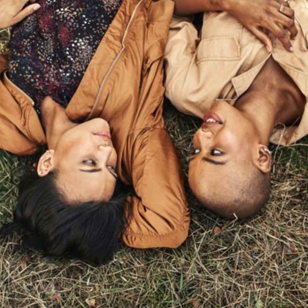 Two women in tan jackets lie head-to-head on grass, one with short hair, the other with long hair, in an outdoor setting.