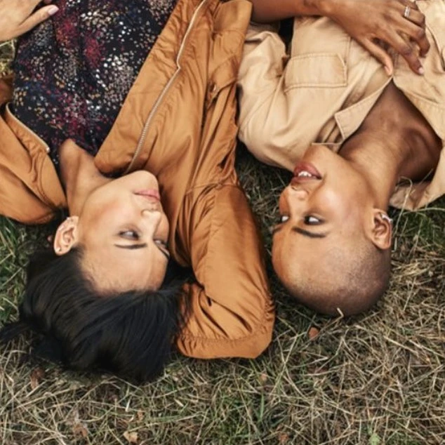 Two women in tan jackets lie head-to-head on grass, one with short hair, the other with long hair, in an outdoor setting.