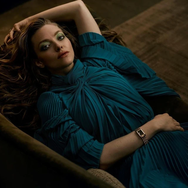 Amanda Seyfried lies on a brown surface wearing a pleated, teal dress and gold watch, with her arm raised above her head.
