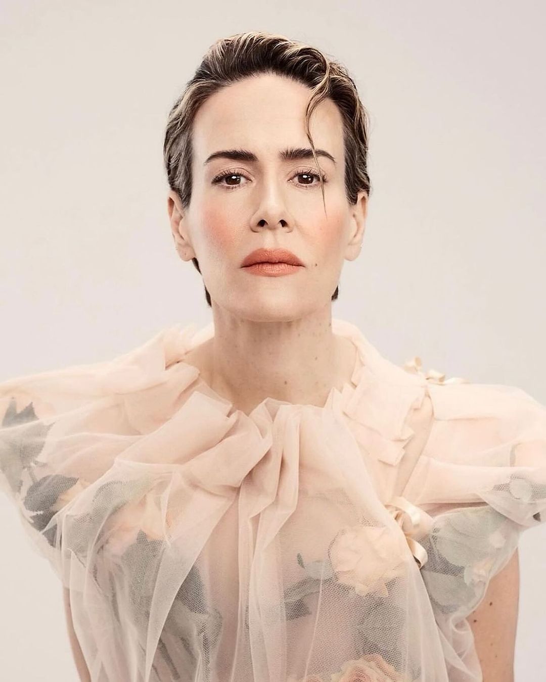 Close-up studio portrait of an adult woman with short hair, wearing a pink ruffled dress, posing against a light backdrop.