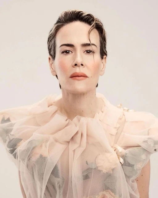 Close-up studio portrait of an adult woman with short hair, wearing a pink ruffled dress, posing against a light backdrop.