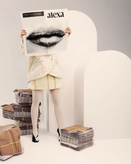 Fashion shot featuring a model holding up a newspaper with a mouth graphic, alongside stacks of newspapers in a clean studio.