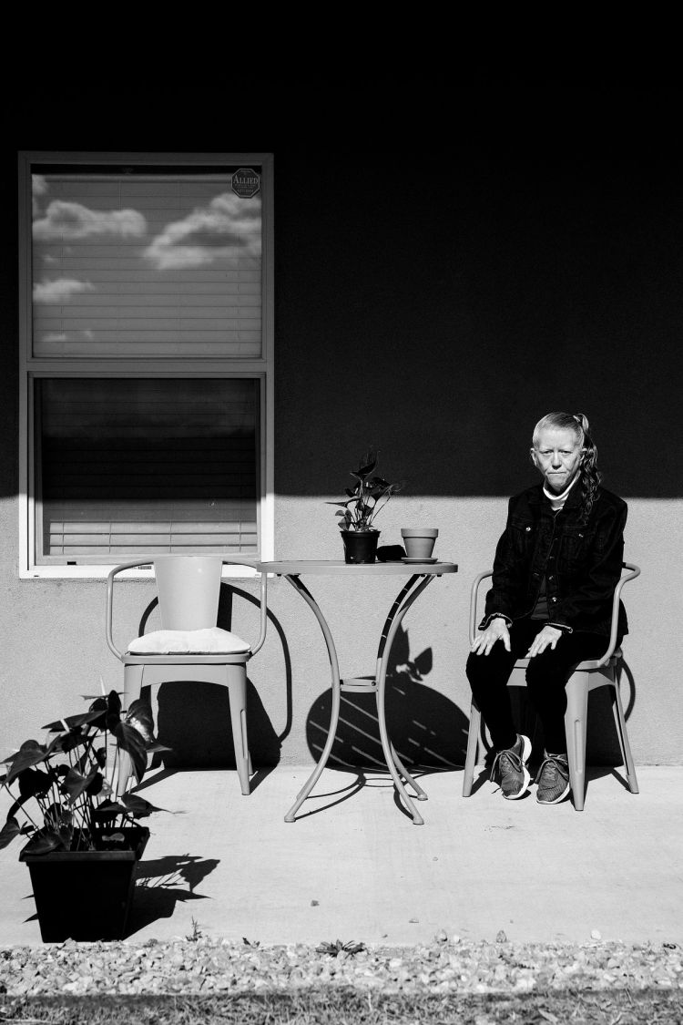 Black and white portrait of a senior woman sitting on a chair on a patio next to a table with potted plants and a window.