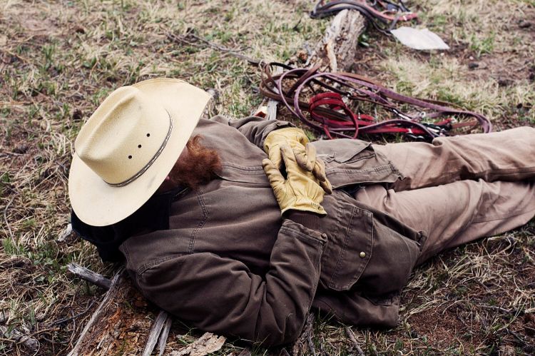 Cowboy lying down on grass with a hat and gloves, wearing a brown jacket and pants. Ropes are lying next to him.