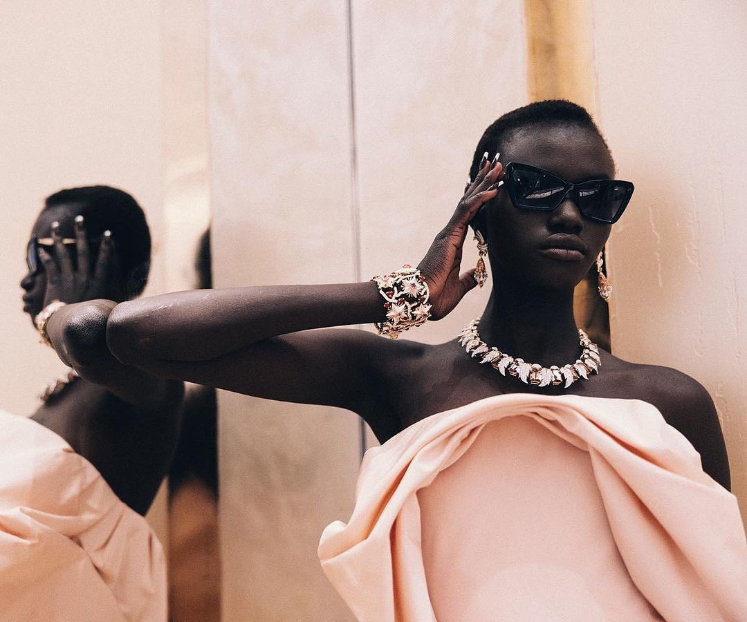 Fashion shot of an African woman in a pink strapless dress wearing sunglasses, a jeweled necklace, earrings, and a bracelet. 