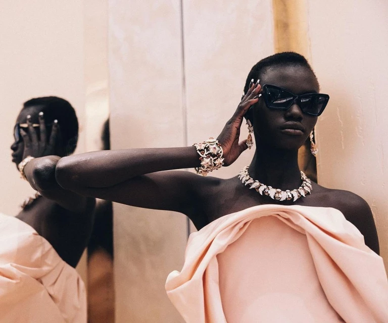 Fashion shot of an African woman in a pink strapless dress wearing sunglasses, a jeweled necklace, earrings, and a bracelet. 