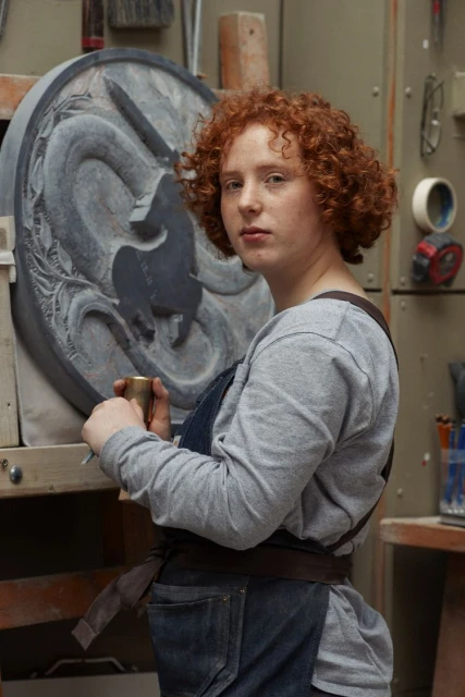 Red-haired adult female artist in an apron stands in a workshop holding a tool, next to a carved stone disc with a dragon design.