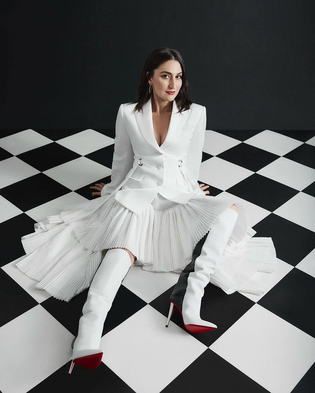 Elegant woman in a white suit and thigh-high boots sits on a black and white checkerboard floor against a black and white wall.