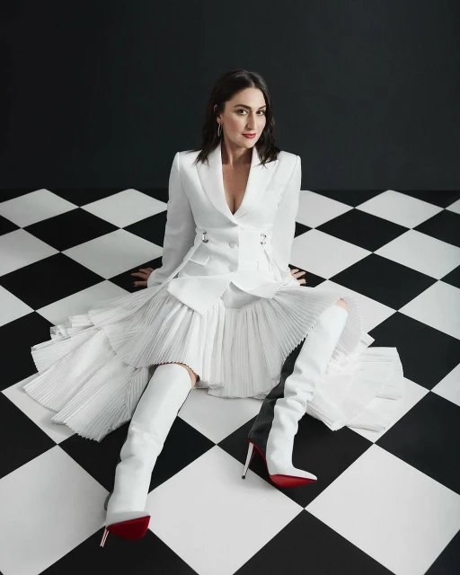 Elegant woman in a white suit and thigh-high boots sits on a black and white checkerboard floor against a black and white wall.