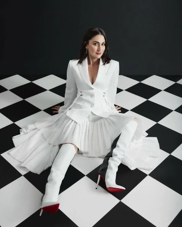 Elegant woman in a white suit and thigh-high boots sits on a black and white checkerboard floor against a black and white wall.
