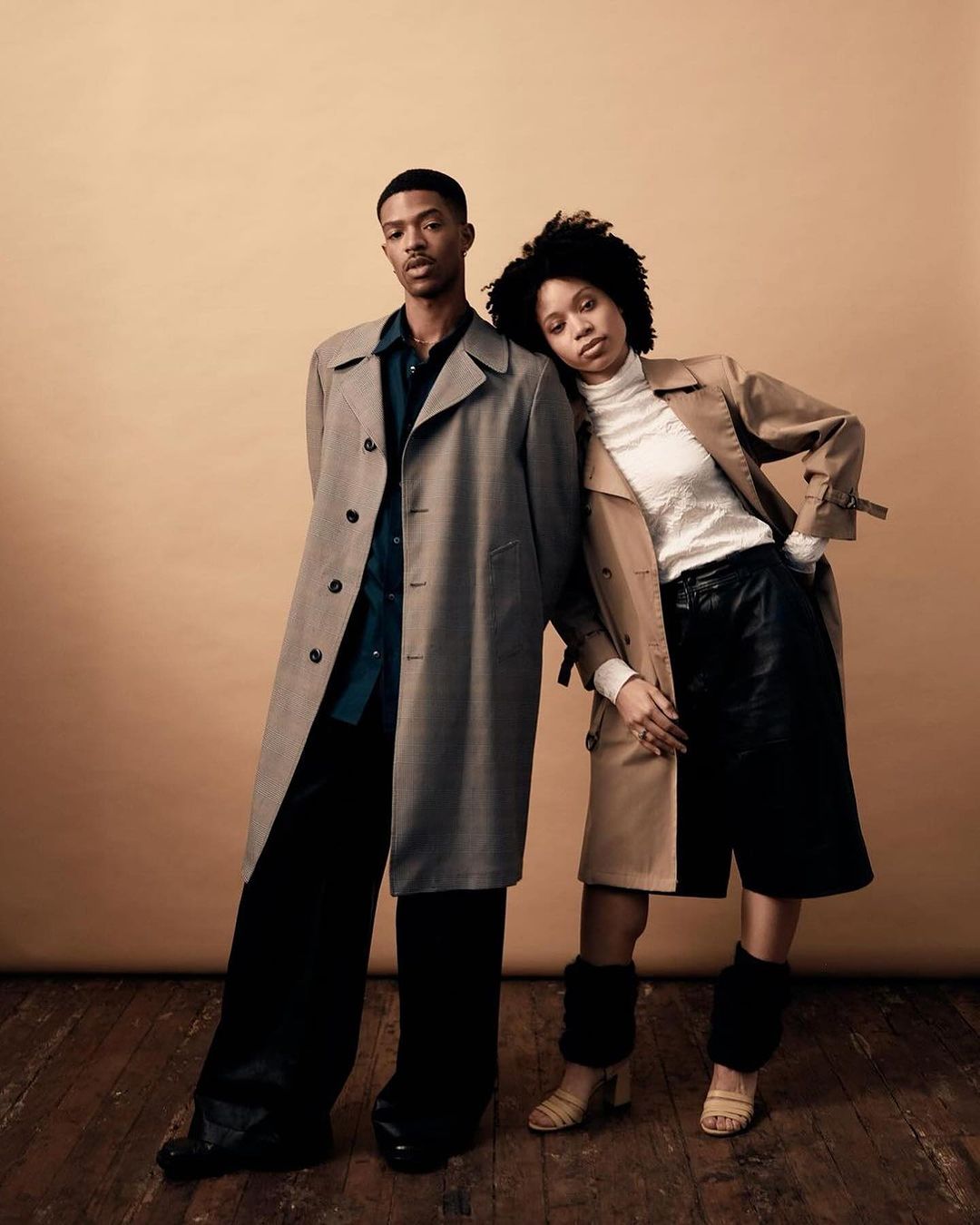Two fashion models wearing trench coats pose in a photo studio against a tan backdrop with a hardwood floor. Editorial fashion photography.