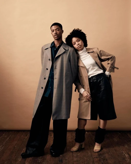 Two fashion models wearing trench coats pose in a photo studio against a tan backdrop with a hardwood floor. Editorial fashion photography.