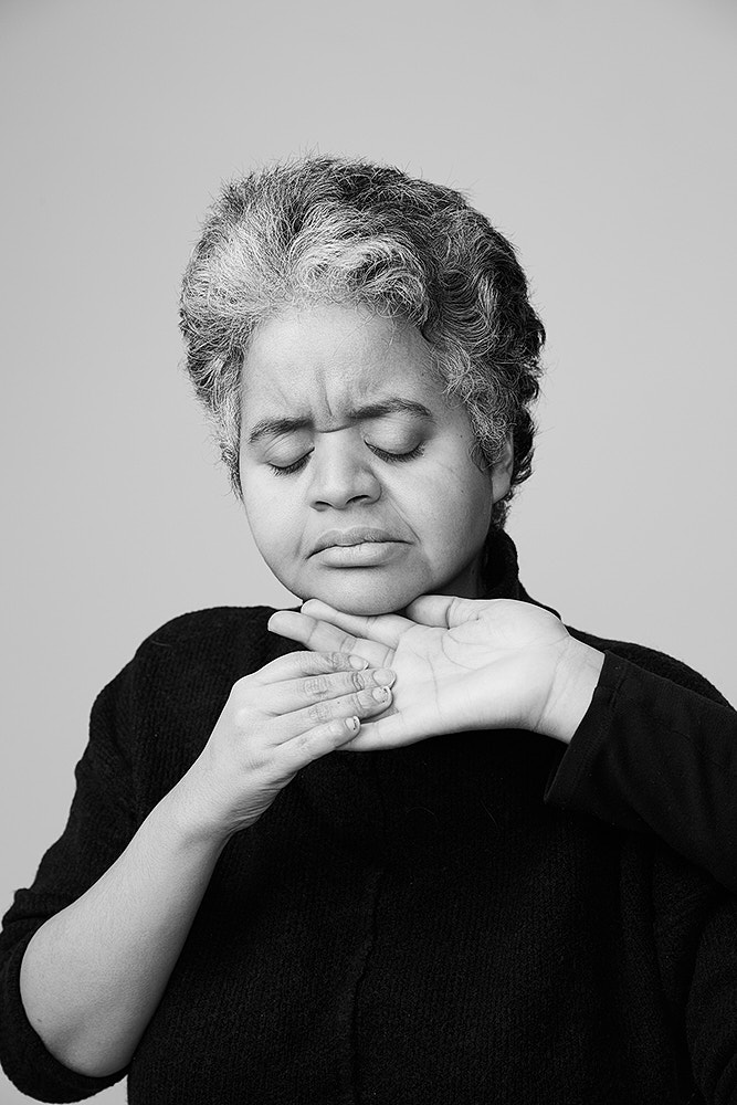 Black and white portrait of an adult woman with her hands on her chin in an indoor studio, captured in eye-level shot.