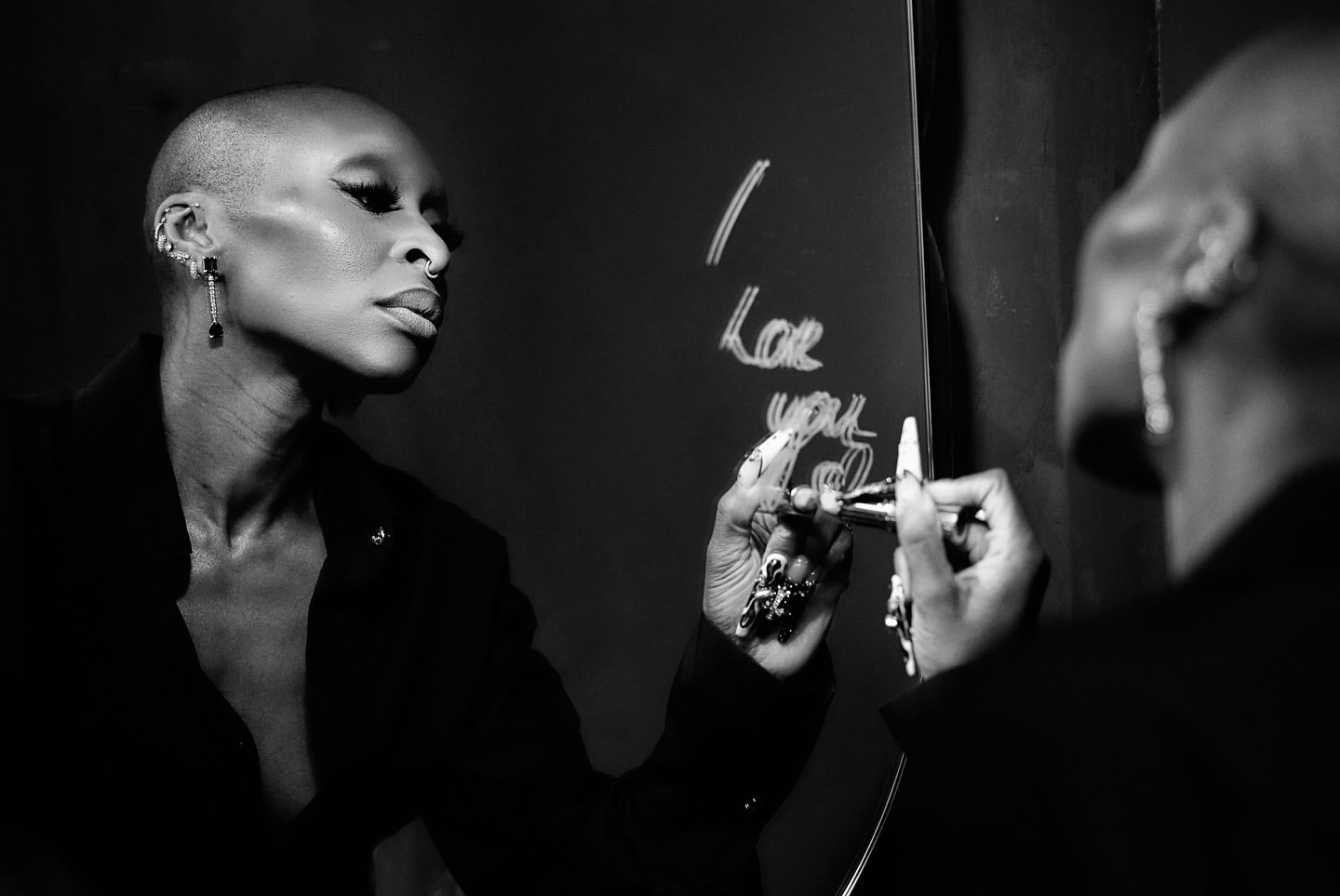 Black-and-white portrait of a bald Black woman with nose ring and earrings writing on a mirror with marker.