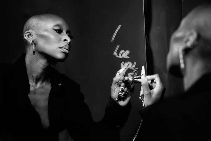 Black-and-white portrait of a bald Black woman with nose ring and earrings writing on a mirror with marker.