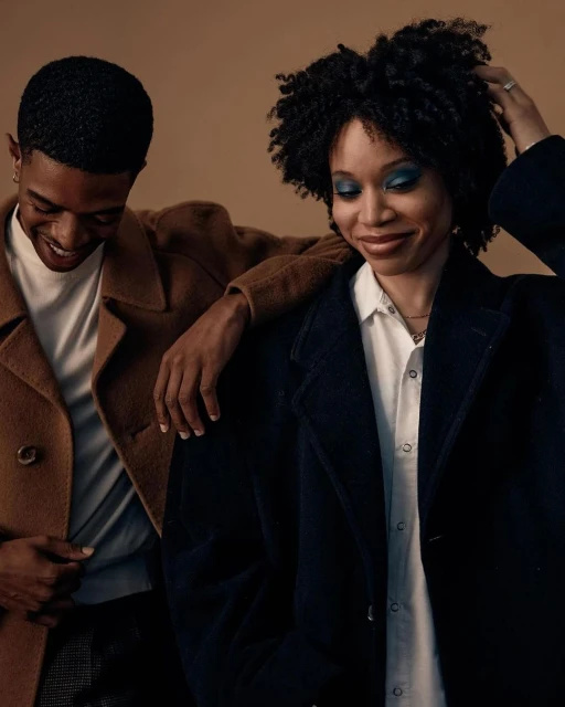 A stylish couple, an adult man and an adult woman, posing indoors wearing fashionable coats against a neutral backdrop, in a portrait composition.