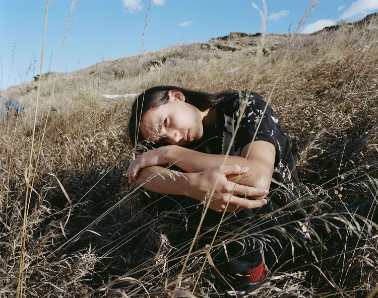 Person with long dark hair crouching amidst dry tall grass under a clear blue sky, photographed by Victoria Will.