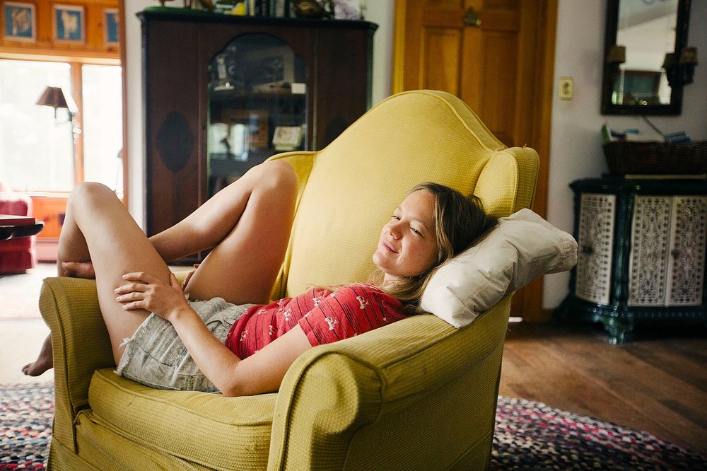 Young woman relaxes barefoot on a yellow armchair with a pillow, wearing a red floral top and grey shorts, with a warm indoor setting.