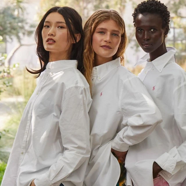 Three women of diverse ethnicities in white collared shirts pose in a bright, airy setting with hints of greenery in the background.