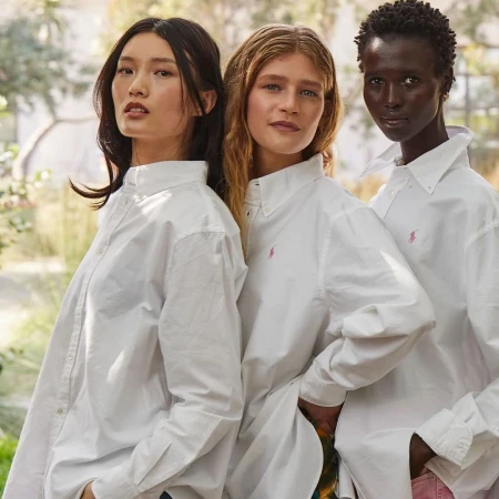 Three women of diverse ethnicities in white collared shirts pose in a bright, airy setting with hints of greenery in the background.
