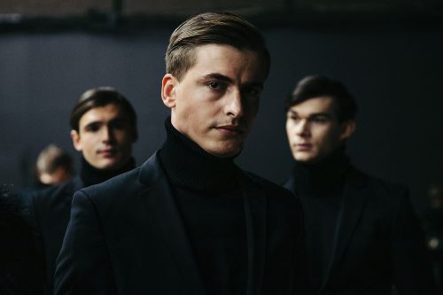 Three adult male fashion models in black turtleneck sweaters and blazers pose indoors against a dark background in a medium shot.